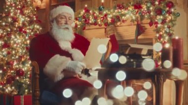 Medium slowmo of big bearded red Santa reading letter from child while sitting by fireplace in beautifully decorated cozy living room