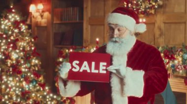 Medium slowmo portrait of surprised Santa in red costume posing for camera with Sale sign in hands in festively decorated living room