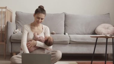 Slowmo of young mother with newborn baby girl sleeping in her arms working on laptop from home, sitting on cozy rug in living room