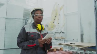 Medium portrait of confident young African American female engineer in safety glasses and hard hat posing for camera with tablet PC while CNC machine working in background at marble processing plant