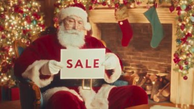 Medium slowmo portrait of kind Santa Claus in red posing for camera with Sale Sign sitting in velvet chair in beautiful living room decorated with Christmas lights