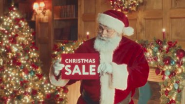 Medium slowmo portrait of bearded Santa smiling at camera holding red Christmas Sale sign on festively decorated background with shining lights