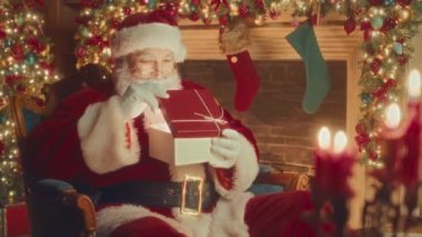 Medium slowmo of delighted Santa in red costume opening Christmas present, sitting in chair by fireplace decorated with sparkling lights