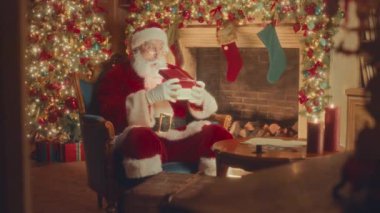 Slowmo of surprised Santa Claus in red costume opening Christmas present box sitting by beautiful fireplace decorated with sparkling lights