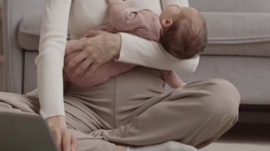 Tilt up slowmo of young Caucasian woman with newborn baby daughter in her arms working on laptop from cozy home while being on maternity leave