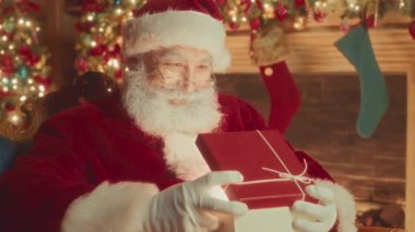Medium slowmo of happy Santa Claus opening Christmas gift box and smiling at camera sitting in his chair by fireplace decorated with sparkling lights