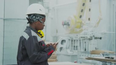 Medium shot of young African American female plant worker in safety glasses and hard hat operating CNC machine with tablet PC