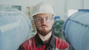 Medium closeup portrait of serious young Caucasian man in safety glasses and hard hat posing for camera standing against large machinery equipment processing natural stone aggregate