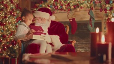 Slowmo of bearded Santa Claus giving Christmas present box to good little African American girl sitting on his laps