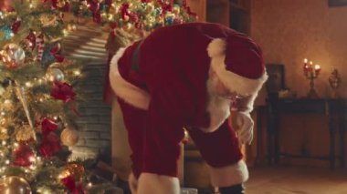 Santa Claus with red bag of Christmas presents sneaking into beautifully decorated house through fireplace and putting boxes under Christmas tree