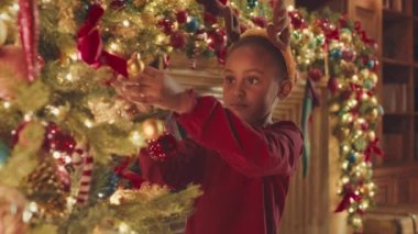 Medium slowmo of pretty little African American girl in reindeer antler headband decorating beautiful Christmas tree on New Year Eve
