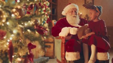 Slowmo of Santa reading Christmas letter of little girl sitting on his laps in cozy living room decorated with sparkly lights
