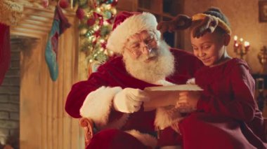 Orta yavaş sakallı Noel Baba, parlak ışıklarla dekore edilmiş güzel oturma odasında, kadife koltukta oturan güzel Afrikalı Amerikalı kızın mektubunu okuyor.