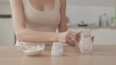 Cropped slowmo of unrecognizable woman making baby formula in milk bottle at kitchen table