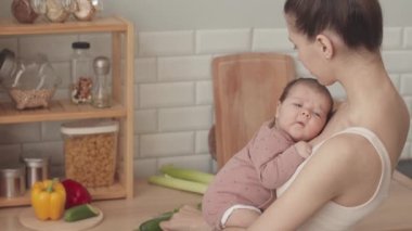 Waist up slowmo of young Caucasian woman with newborn baby daughter in her arms making healthy breakfast in minimalist kitchen