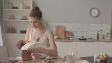 Medium slowmo of young Caucasian woman with newborn baby in her arms working from home, sitting at kitchen table in light cozy apartment
