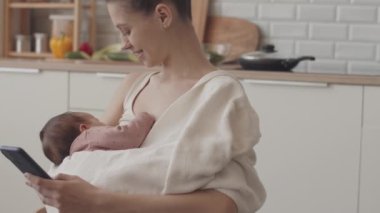 Tilt up slowmo of young smiling woman using her smartphone while holding her newborn baby in hands sitting at cozy kitchen table
