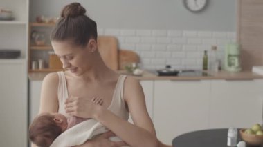 Medium slowmo portrait of young beautiful Caucasian woman posing for camera at cozy kitchen holding her newborn baby daughter in her arms