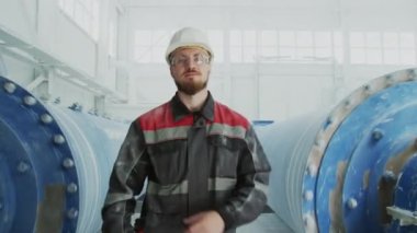 Medium portrait of confident young male plant worker in uniform posing with hands folded against two grinding machines processing marble aggregate used in construction