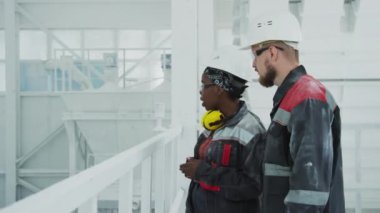 Medium shot of young African American female plant worker explaining duties to new colleague, looking together at modern machinery equipment processing natural stone