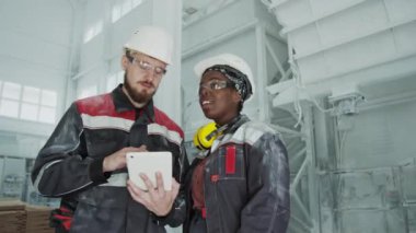 Medium shot of multiethnic couple of engineers in uniforms and hard hats working together at stone granite factory with modern equipment and high quality raw materials