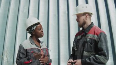 Medium shot of two multiethnic factory workers in uniforms and hard hats having conversation outdoors