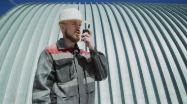 Medium shot of Caucasian foreman in uniform and hard hat speaking through walkie talkie outdoors