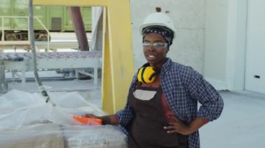 Medium portrait of confident African American female marble production factory worker posing for camera with stone slabs on crane outside plant
