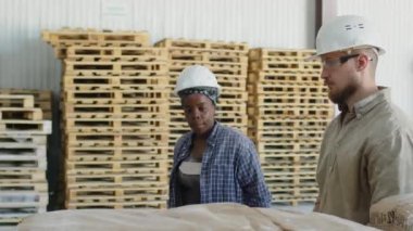 Medium shot of diverse couple of coworkers talking while walking along aisles with pallets at warehouse