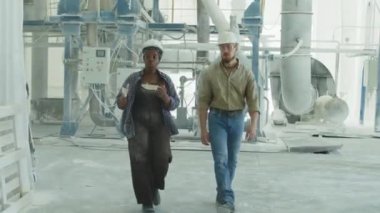 Full length tracking shot of multiethnic couple of coworkers talking while walking along industrial plant processing cement