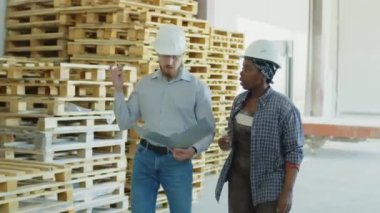 Medium shot of foreman talking to African American female employee while walking along wooden pallets at warehouse