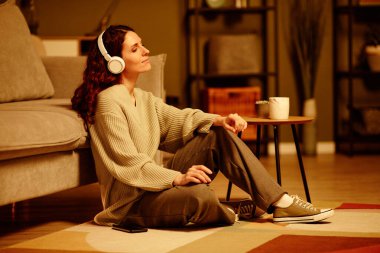 Gündelik giysiler giyen şık beyaz kadın oturma odasında rahat rahat oturup akşamları kablosuz kulaklıkla müzik dinliyor.