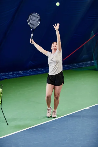 屋内裁判所でテニスをしている若い白人女性の垂直肖像画と空気中のボールを投げる — ストック写真