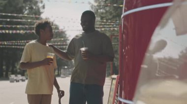 Orta boy Afro-Amerikan bir adam ve 12 yaşındaki oğlunun sokak yemekleri kamyonunda içki alırken sohbet ederken ve iyi vakit geçirirken, güneşli bir günde hafta sonlarını parkta birlikte geçirirken.