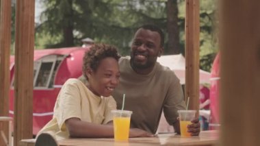 Neşeli siyahi bir adam ve 12 yaşındaki oğlu parkta bir kafede oturmuş portakal suyu içiyor, sohbet ediyor ve eğleniyorlar.
