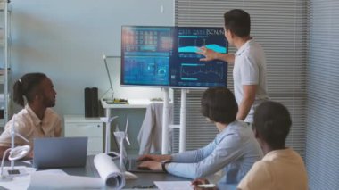 Genç Asyalı erkek mühendis projektör ekranında çizelgeler ve kağıt üzerinde planlar gösterirken aynı zamanda sürdürülebilirlik projesini ofisteki çeşitli meslektaşlarına sunuyor.