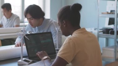 Genç Afrikalı Amerikalı kadın mühendis, çeşitli meslektaşlarıyla ofiste dizüstü bilgisayar üzerinde çalışırken teknik planları inceliyor.