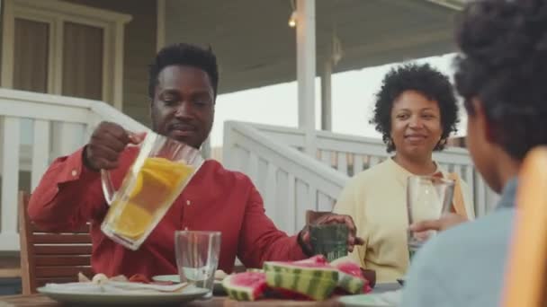 陽気なアフリカ系アメリカ人の家族が 晴れた日の裏庭で友人たちと昼食をとる中型ショット テーブルでゲストに自家製レモネードを注ぐ黒人男性 — ストック動画