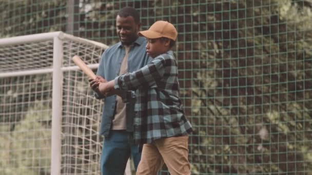 アフリカ系アメリカ人の男が 公園の草の上の野外で一緒に練習しながら野球でバットする方法を示すスローモードを傾けました — ストック動画
