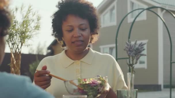 興奮した黒人女性が裏庭の緑の芝生に家族と屋外の夏の夕食をしながらプレートに新鮮な健康的なサラダを置くのを待つ — ストック動画