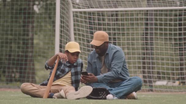 アフリカ系アメリカ人の父親と10代の息子のスローモーは 野球トレーニング後にスポーツフィールドで緑の草の上に休んでいる間 スマートフォンでセルフィーの肖像画を撮ります — ストック動画