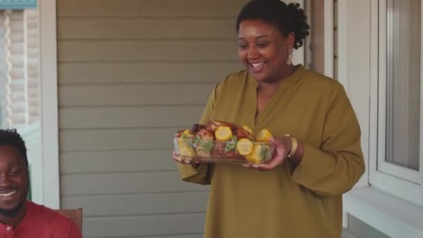 ワクワクする若い黒人女性のミディアムショットは 家族と一緒に家のポーチで夕食をしながらテーブルにジャガイモで美味しいチキンを提供しています — ストック動画