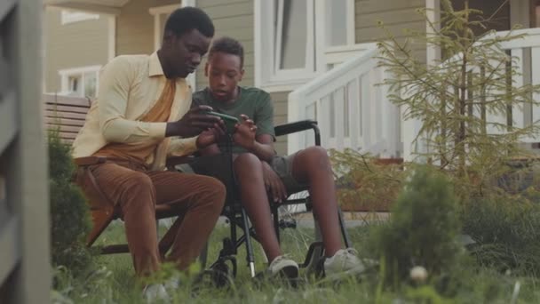 12歳のアフリカ系アメリカ人の男の子は 暖かい晴れた日に家の外の庭で屋外で過ごしながら 兄とスマートフォンでゲームをする — ストック動画
