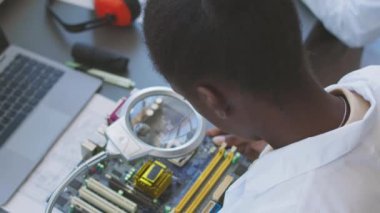 Laboratuvarda araştırma yaparken PCB 'yi büyüteçle inceleyen Afrikalı Amerikalı bilgisayar mühendisliği öğrencisi.