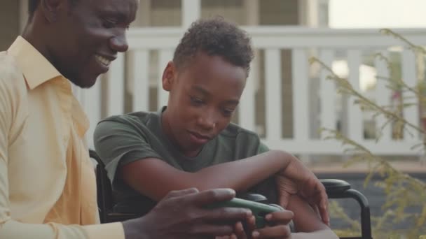 アフリカ系アメリカ人の若者と彼の小さな車椅子の兄弟は家の外で一緒に時間を過ごし スマートフォンでゲームをする — ストック動画