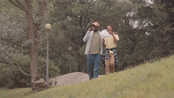 夏の日に公園で木々の間を歩いている間 十代の息子と黒人の男の全長の遅いスローモ — ストック動画