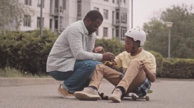 Slowmo of caring African American father attaching elbow pads on his son arms while teaching him skateboarding outdoors in park