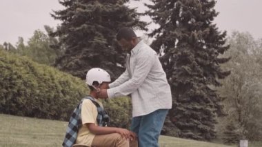 Slowmo of Black man putting on helmet on head of his teenage son while teaching him to ride skateboard outdoors in park