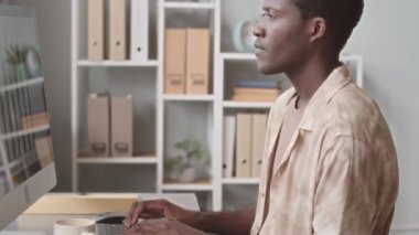 Side view tilt up slowmo of young Black man smoking electronic cigarette while working with business graphics on computer in office