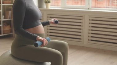 Cropped slowmo of unrecognizable pregnant woman in activewear sitting on fit ball and lifting dumbbells